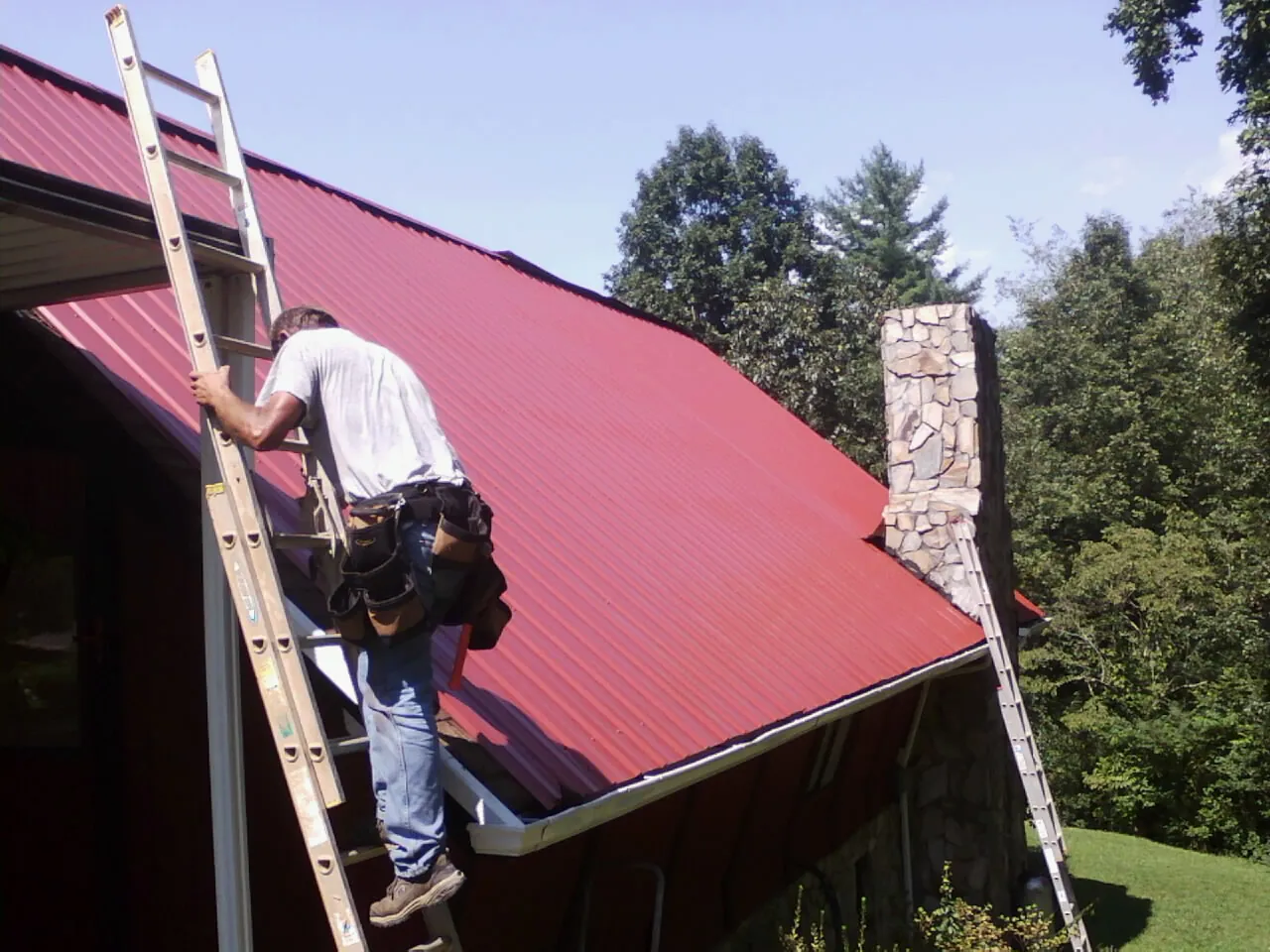 Roofer on a ladder beside a red metal roof and stone chimney — Soffit & Fascia Repair by Roofing Brothers in Livingston